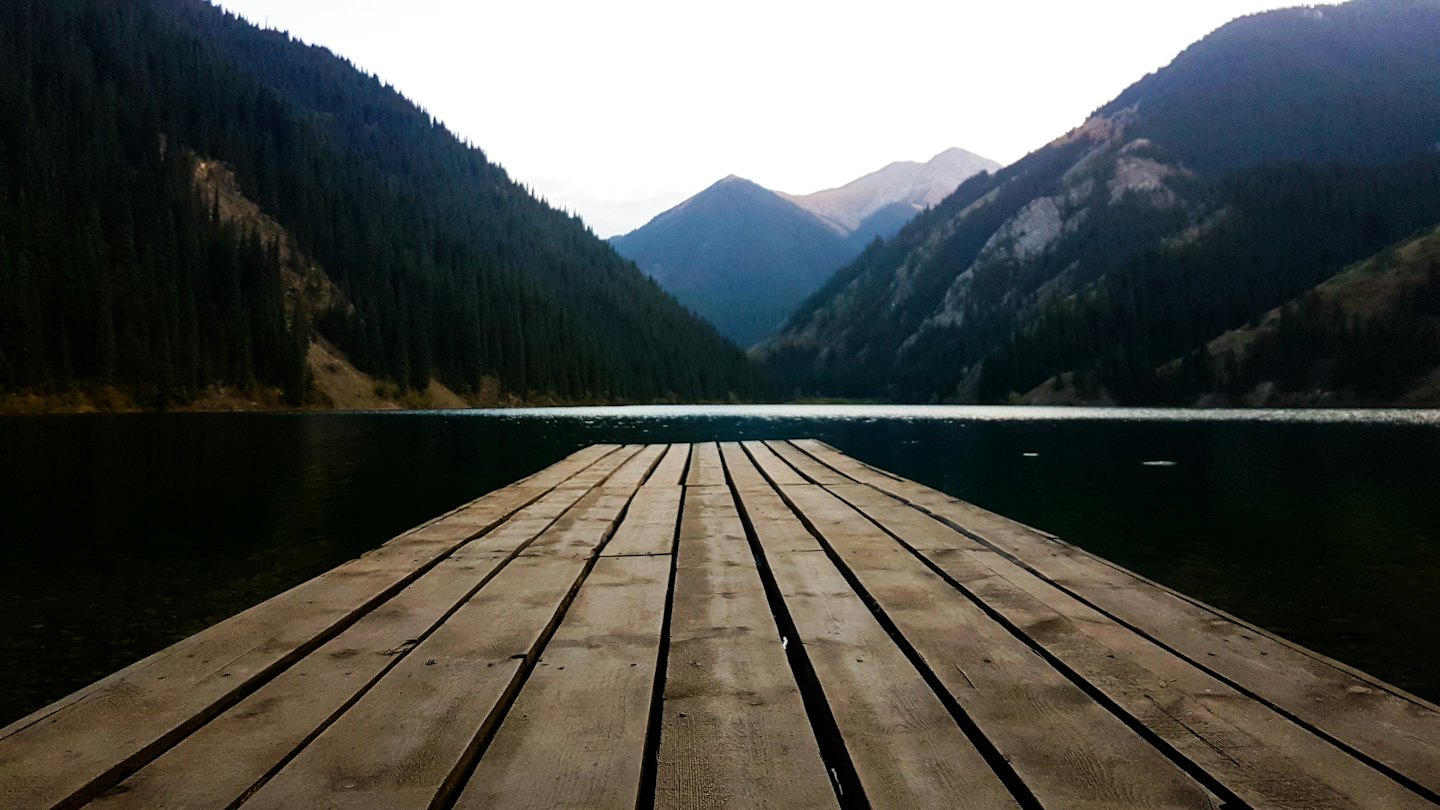 A long wooden dock extends into a lake surrounded by mountains.