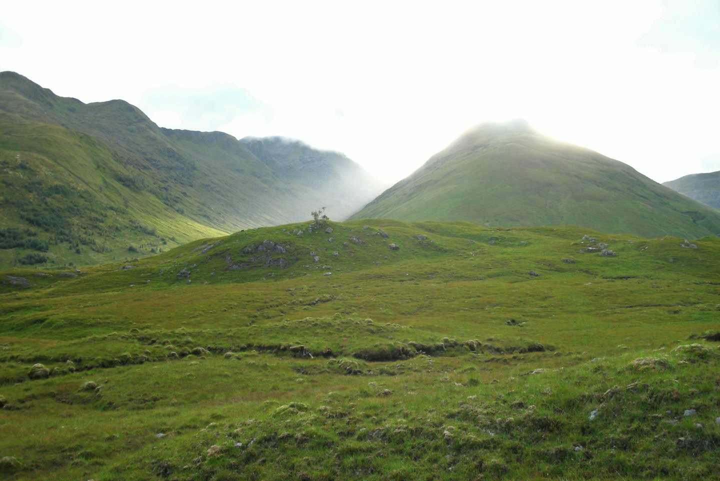 Sunlight breaks through mist over rolling green hills and mountains.
